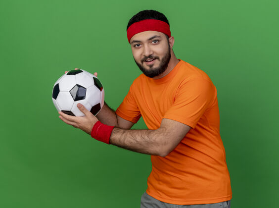 年轻戴着头带和腕带的年轻运动型男子高兴地在绿色背景上孤立的一侧举着球请壁板运动
