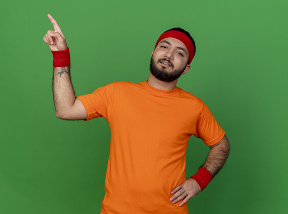 头带自信的年轻运动型男子戴着头带和腕带 将手放在臀部 并在绿色背景上孤立地指向侧面 留有复制空间壁板信心运动