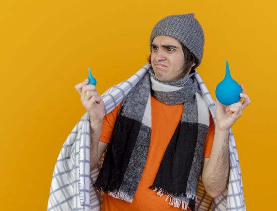 年轻一个戴着冬天帽子 披着方格围巾 拿着东西看橙色背景上孤立的灌肠的年轻病患抱着疾病围巾