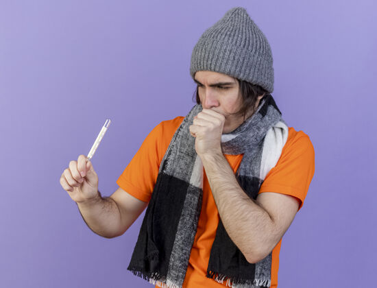 年轻人咳嗽的年轻病人戴着冬天的帽子 戴着围巾 看着隔离在紫色背景上的温度计生病围巾体温计