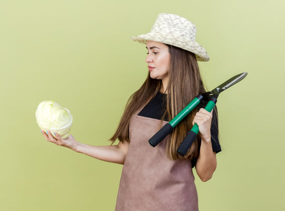 思考想着穿着制服的漂亮园丁女孩戴着园艺帽把剪刀放在肩上 看着她手上的卷心菜孤立在橄榄绿的背景上帽子剪子肩膀