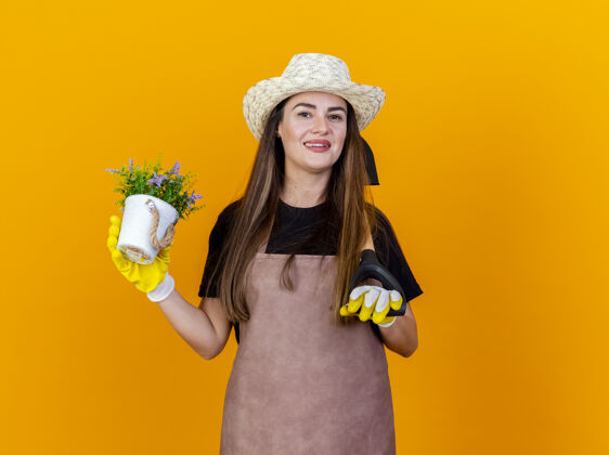 花盆微笑美丽的园丁女孩穿着制服 戴着园艺帽 戴着手套 肩上扛着铁锹 把花放在橙色背景的花盆里微笑帽子女孩