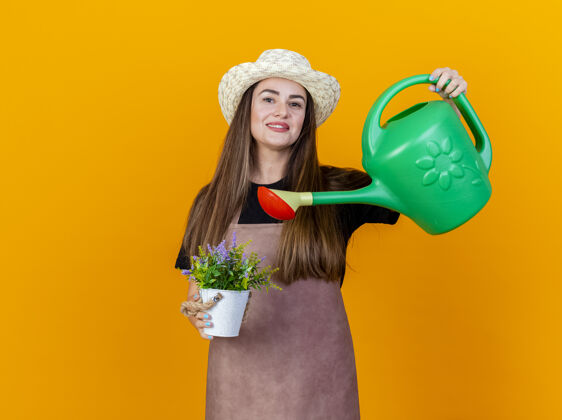 女孩微笑美丽的园丁女孩穿着制服 戴着园艺帽 在花盆里浇花 可以在橙色的背景上隔离花浇水园艺