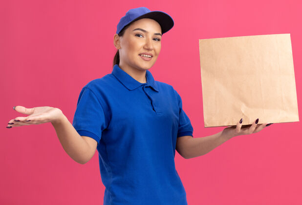 包装身穿蓝色制服 头戴鸭舌帽的年轻女送货员拿着纸包 面带微笑地站在粉色的墙上 脸上洋溢着幸福的表情年轻微笑快乐