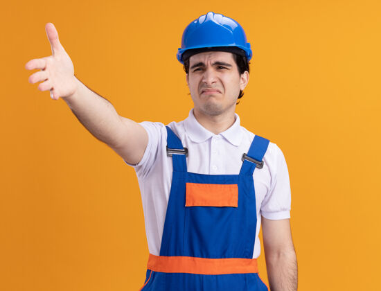 愤怒年轻的建筑工人穿着建筑制服 戴着安全帽 看上去很困惑 站在橙色的墙上 愤怒地举起手来站立年轻安全