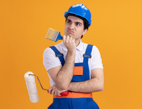靠边年轻的建筑工人穿着建筑制服 戴着安全帽 手里拿着油漆刷和滚筒 站在橙色的墙上困惑地看着一边建筑工人头盔滚轴