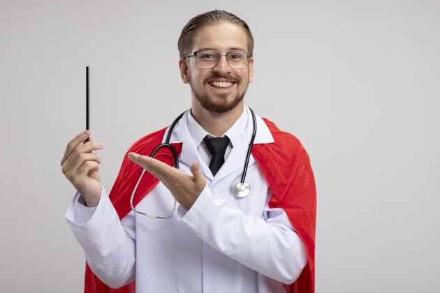 手微笑着的年轻超级英雄 穿着医用长袍 手持听诊器和眼镜 用手指着白色背景上的铅笔听诊器年轻铅笔
