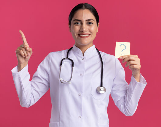 提醒快乐的年轻女医生 身穿医用外套 手持听诊器 手持问号提示纸 食指朝上 站在粉红色的墙上 愉快地微笑着外套指着举行