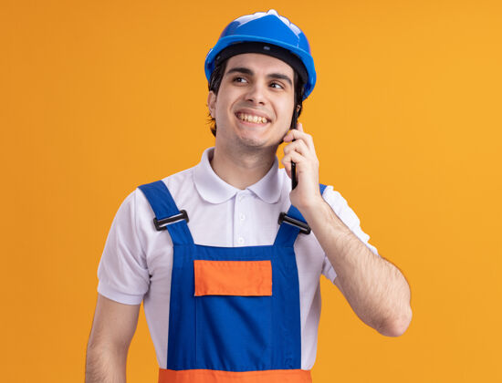 手机身穿施工制服 头戴安全帽的年轻建筑工人站在橙色的墙上 面带微笑地一边看一边讲着手机建筑工人安全男人