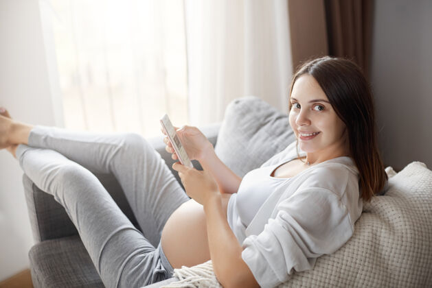 期望年轻的孕妇使用智能手机呆在家里的照片和她的朋友在线聊天女孩微笑年轻