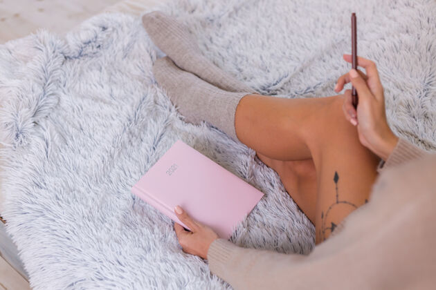 计划者穿着羊毛袜子和毛衣的女人 粉色笔记本上写着2021 臀部有大纹身女人坐在卧室的家里的床上坐着早晨袜子