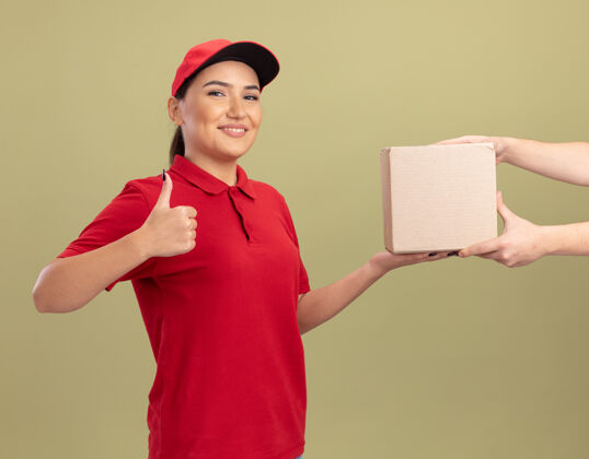 拇指年轻的送货员身着红色制服 戴着帽子 站在绿色的墙上 友好地微笑着竖起大拇指 把纸板箱递给顾客制服顾客站立