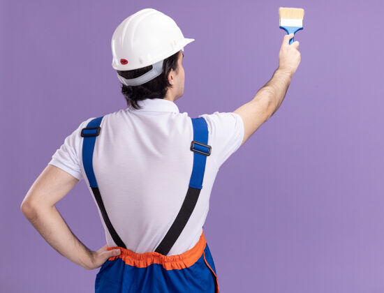 年轻年轻的建筑工人穿着建筑制服 戴着安全帽 背着油漆墙站着 油漆辊压在紫色的墙上站头盔滚轴