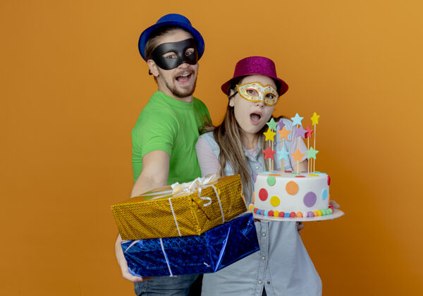 化装舞会惊讶的年轻女孩戴着粉色帽子和化装眼罩拿着生日蛋糕 快乐的帅哥戴着蓝色帽子戴着化装眼罩拿着礼盒橙色男人帅哥