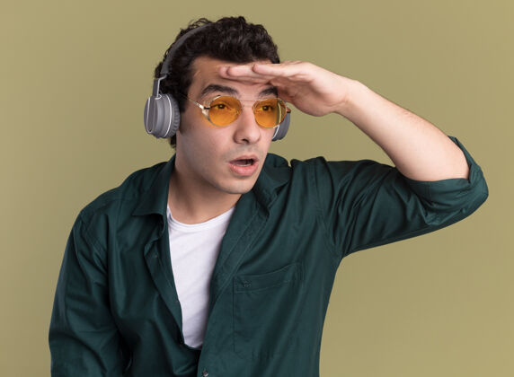 男人穿绿衬衫的年轻人戴着眼镜 戴着耳机 手举着头远远地站在绿墙上衬衫走着手