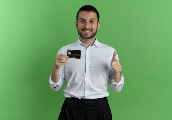商人笑容可掬的帅哥拿着信用卡 竖起大拇指孤零零地站在绿色的墙上人优雅拇指