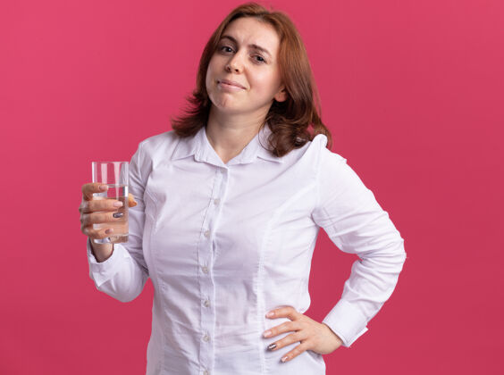 杯子身穿白衬衫的年轻女子举着一杯水 站在粉红色的墙上 面带微笑地看着前面抱着衬衫站着