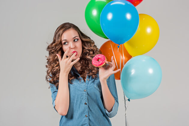 成人一个年轻的女人在灰色的工作室墙上吃着甜甜圈 手里拿着五颜六色的气球华丽年轻魅力