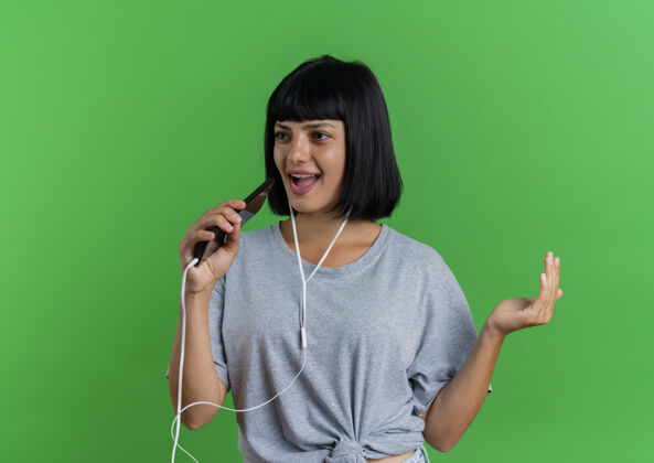 黑发戴着耳机的快乐的年轻黑发白种女人拿着电话假装在绿色背景上唱歌 并留有复制空间耳机背景电话