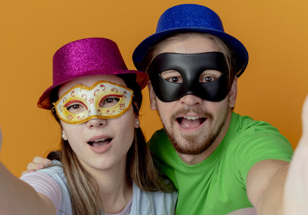 情侣一对年轻快乐的夫妇戴着粉色和蓝色的帽子 戴上化装眼罩 假装站在橙色的墙上 看起来与世隔绝橙色粉色举行
