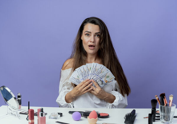 工具惊讶的漂亮女孩坐在桌子旁 手里拿着化妆工具 手里拿着隔离在紫色墙上的钱紫色化妆美女