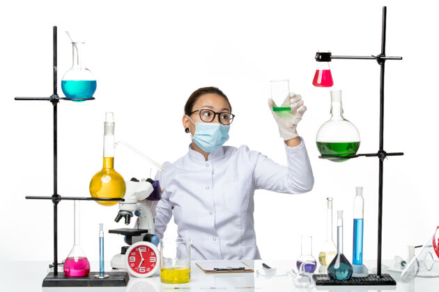 不同前视图穿着医疗服的女化学家 戴着面罩 在浅白色背景上拿着不同的溶液 飞溅着病毒化学实验室的病毒视图药品实验室