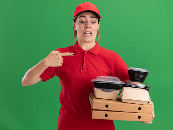 盒子焦急的年轻漂亮的女送货员穿着制服 拿着并指着绿色比萨饼盒上的纸食品包装和容器点姿势人