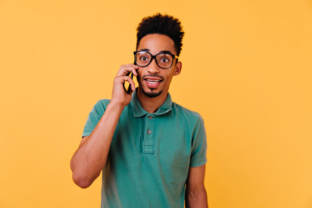 沟通一个戴着大眼镜的男孩在打电话一个穿着绿色t恤衫的情绪激动的非洲男人在打电话给别人的室内肖像休息手机乐趣