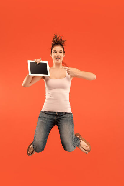 成人蓝色背景下的年轻女子在跳跃时使用笔记本电脑或平板电脑的图像完整持有科技