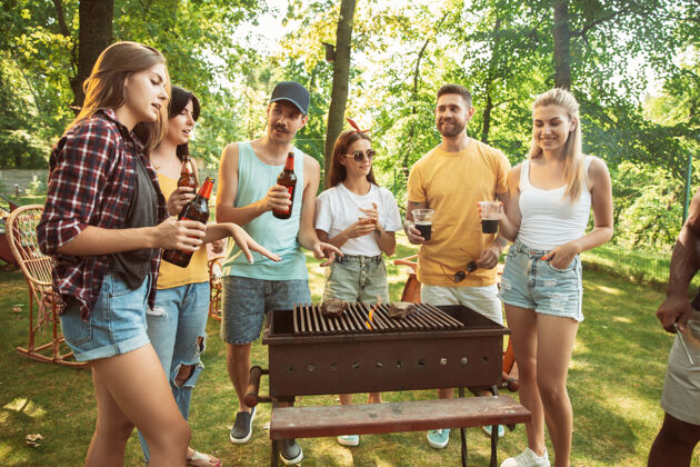 享受一群快乐的朋友在阳光明媚的日子里举行啤酒和烧烤派对一起在户外的森林空地或后院休息欢庆和放松 欢笑夏日生活方式 友谊理念家庭烧烤啤酒
