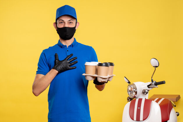 工作正面图：戴着口罩的男快递员在黄色大流行的快递工作服上送咖啡男性大流行护目镜