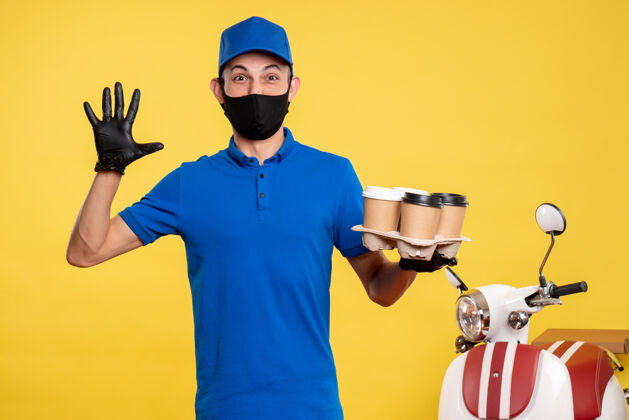 男性正面图：戴着黑色口罩的男信使拿着咖啡 戴着黄色工作帽-大流行快递工作服务工作商务持有