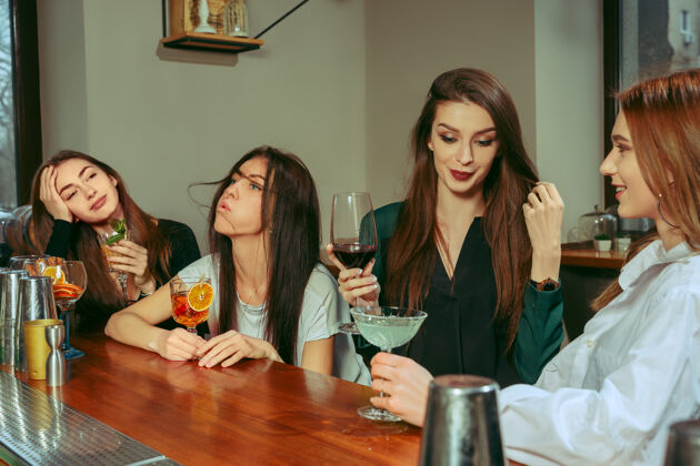 酒吧女朋友在酒吧喝酒她们坐在一张摆着鸡尾酒的木桌旁她们穿着休闲服手白天夜晚