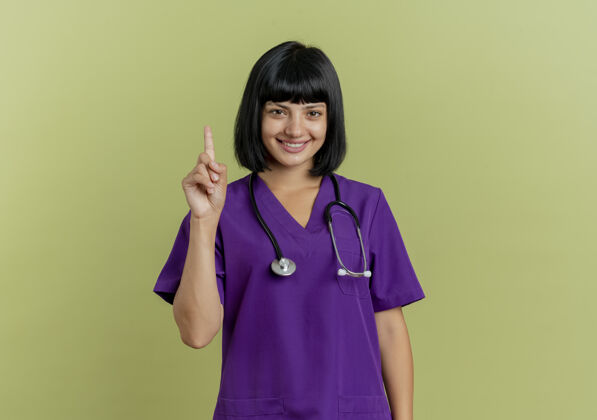 黑发微笑着的年轻黑发女医生穿着制服 手持听诊器 抬头看着隔离在橄榄绿背景和复印空间上的摄像头背景年轻微笑