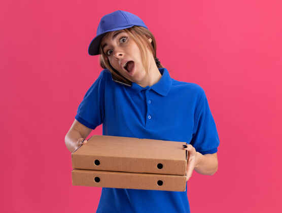 盒子穿着制服的年轻漂亮的送货女孩拿着比萨饼盒 在粉红色的桌子上打电话年轻漂亮披萨