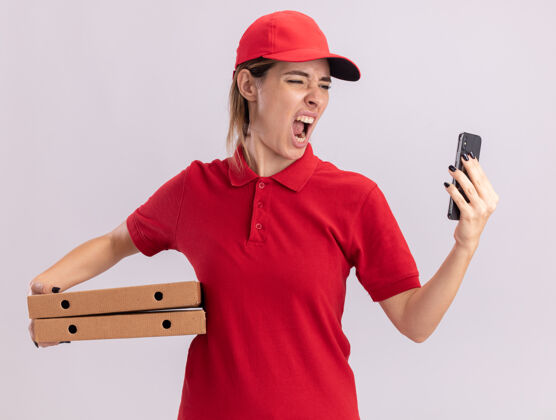 年轻穿着制服的愤怒的年轻漂亮送货女孩拿着比萨饼盒 看着白色的手机漂亮人披萨
