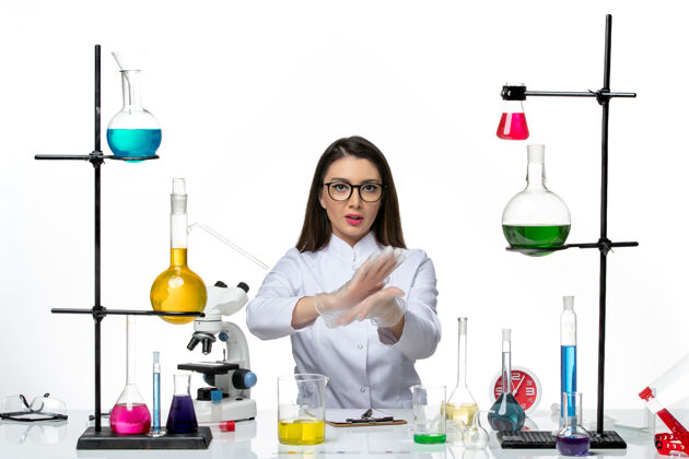 坐着前视图：穿着白色医疗服的女化学家正坐在白色办公桌上 拿着解决方案——科学大流行实验室病毒瓶子前面液体