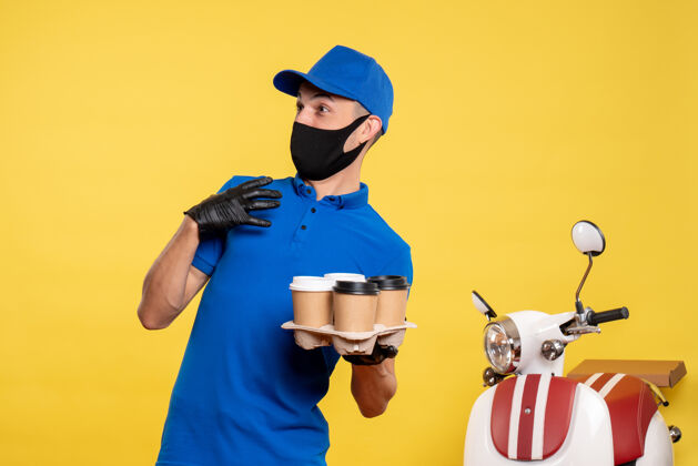 咖啡正面图：戴着黑色口罩的男快递员拿着咖啡在黄色大流行的快递工作服上工作工作前面流行病
