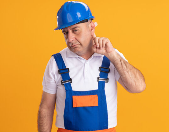 人无知的白人成年建筑工人穿着制服 把手指放在耳朵后面放在桔子上男人制服无知