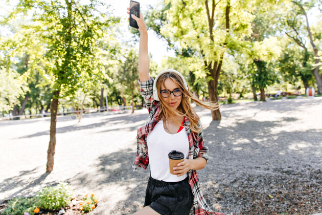 青少年悠闲的女人端着一杯咖啡在公园里翩翩起舞温文尔雅的金发女郎戴着眼镜 在早晨欣赏大自然的音乐人欢笑休闲