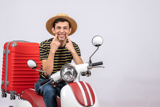 运动正面图：骑着轻便摩托车戴着草帽的快乐的年轻人指着他的微笑坐着视图帽子