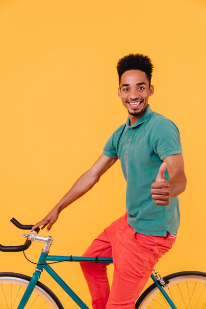 享受高兴的非洲自行车手摆姿势 拇指向上室内拍摄的黑色男子在休闲绿色t恤坐在自行车上现代模特表情