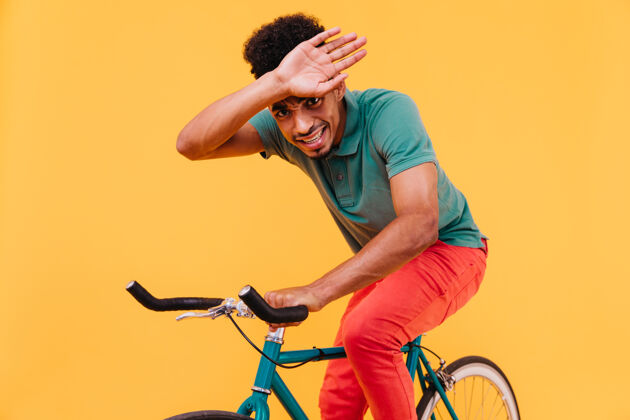 帅气穿着鲜艳衣服的幽默男模特在自行车上摆姿势热情的黑人年轻人坐在绿色自行车上鬼混的室内照片黑色微笑现代
