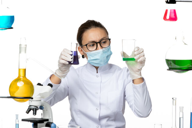 持有前视图穿着医疗服的女化学家 戴着面罩 在白色背景上拿着不同的溶液 飞溅着病毒化学实验室的病毒病毒正面解决方案