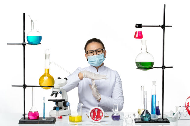 坐前视图穿着白色医疗服的女化学家带着面罩坐在浅白背景上的溶液化学家病毒-飞溅实验室专业实验室前面