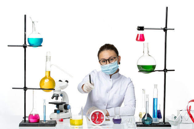 实验室前视图穿着医疗服的女化学家戴着面罩坐着 拿着溶液 在浅白色背景上写笔记化学家实验室病毒covid-飞溅书写前面套装