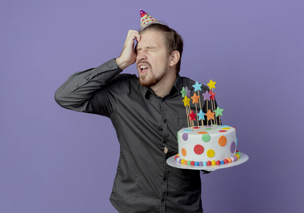 帅气恼怒的帅哥戴着生日帽把手放在额头上 把蛋糕隔离在紫色的墙上手生日额头
