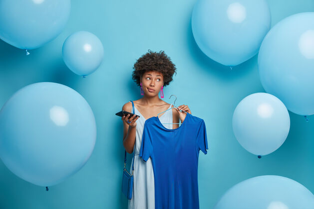 氦体贴的黑皮肤年轻女子卷发 衣架上拿着优雅的蓝色连衣裙 手里拿着手机 穿着蓝色主题派对的衣服 一边看 对着气球摆出沉思的样子衣柜女人装置