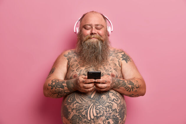 电话满意的成年胖男室内镜头手持手机 使用音乐应用程序 耳朵戴立体声耳机 听歌曲 赤身裸体 有纹身 隔离在粉色墙上超重成人罗西快乐