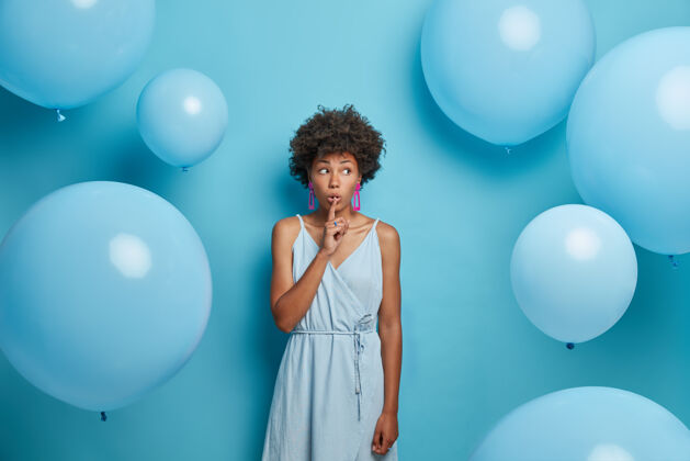 手指非裔美国女士的室内镜头将食指按在嘴唇上 显示出安静的标志 说出秘密 散布谣言和旁观者 在社交活动上的空闲时间穿着裙子 一切都是蓝色的年轻安静惊喜